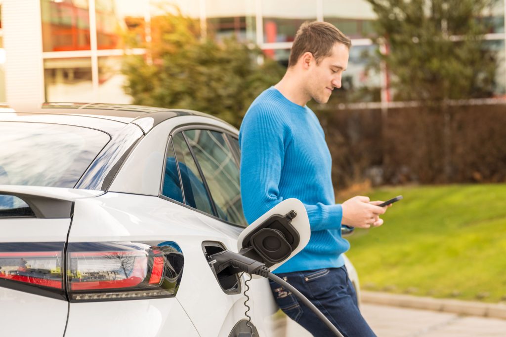 Mann, der an einem E-Auto lehnt und Handy schaut während das Auto aufgeladen wird.